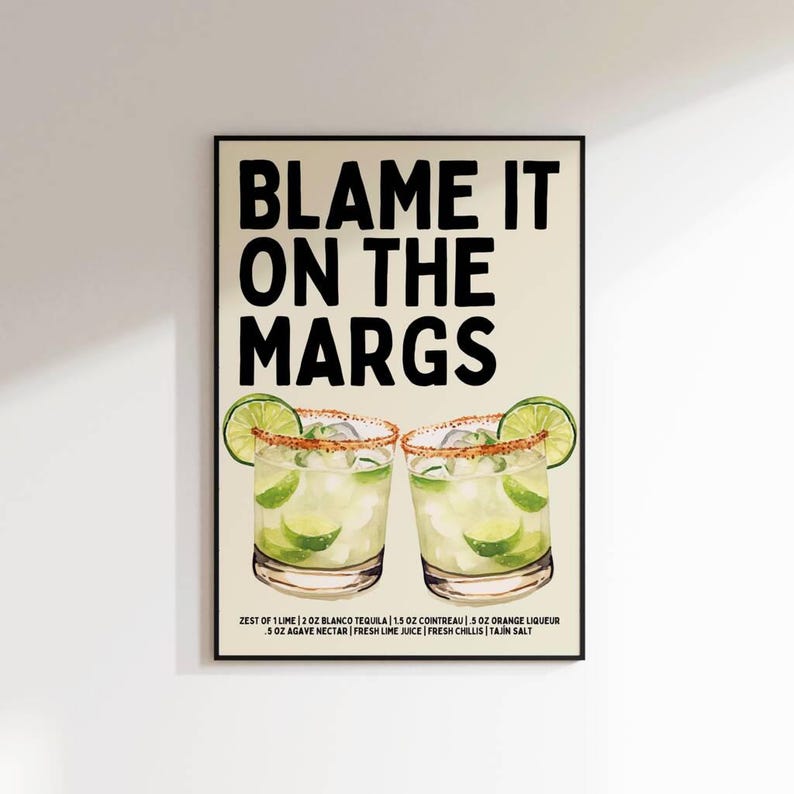 May include: A framed art print with the text "BLAME IT ON THE MARGS" in bold black letters. Below the text are two margarita cocktails with lime wedges and a recipe.