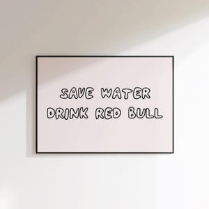 May include: A framed print with the text "SAVE WATER DRINK RED BULL" in black, handwritten-style lettering on a light pink background. The frame is black, and the print is displayed against a white wall.