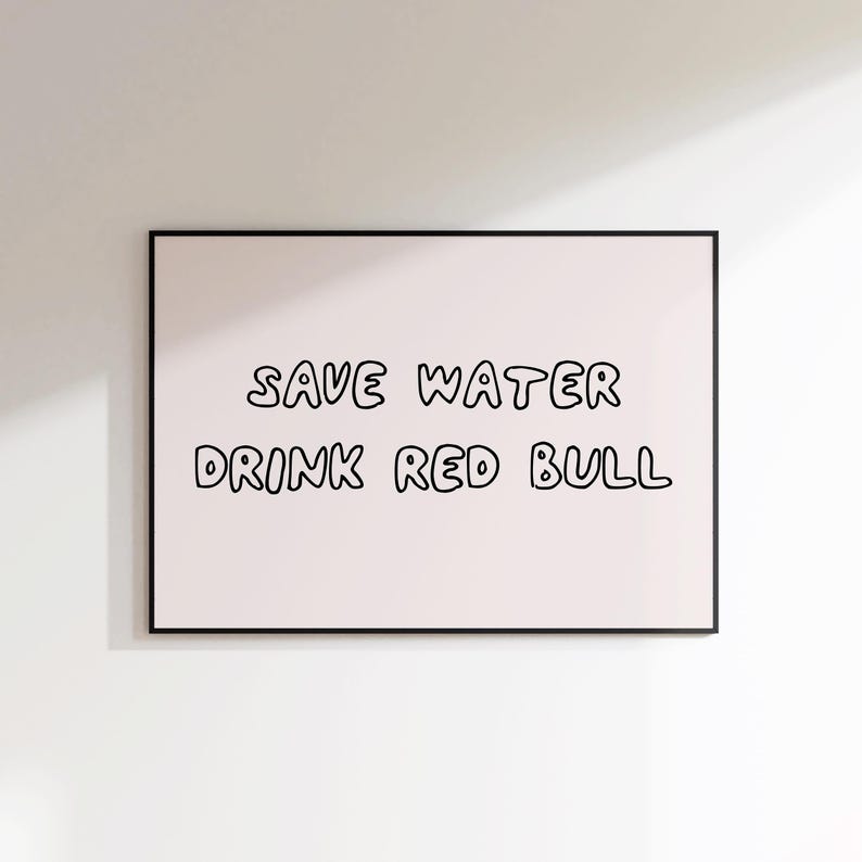 May include: A framed print with the text "SAVE WATER DRINK RED BULL" in black, handwritten-style lettering on a light pink background. The frame is black, and the print is displayed against a white wall.