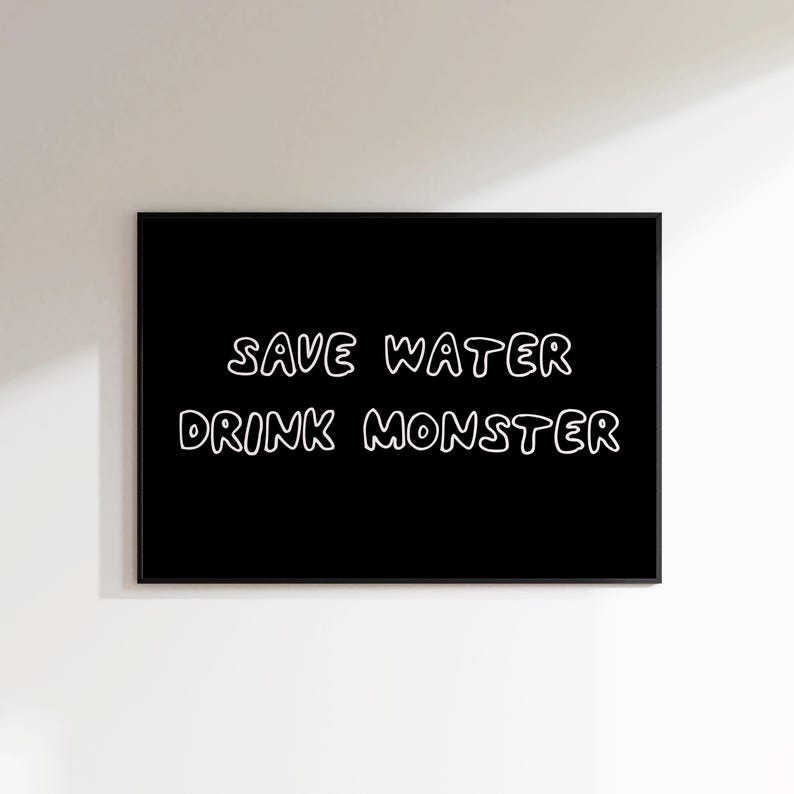 May include: A black framed art print with white text that reads "SAVE WATER DRINK MONSTER". The text has a handwritten style. The art print is hanging on a white wall.