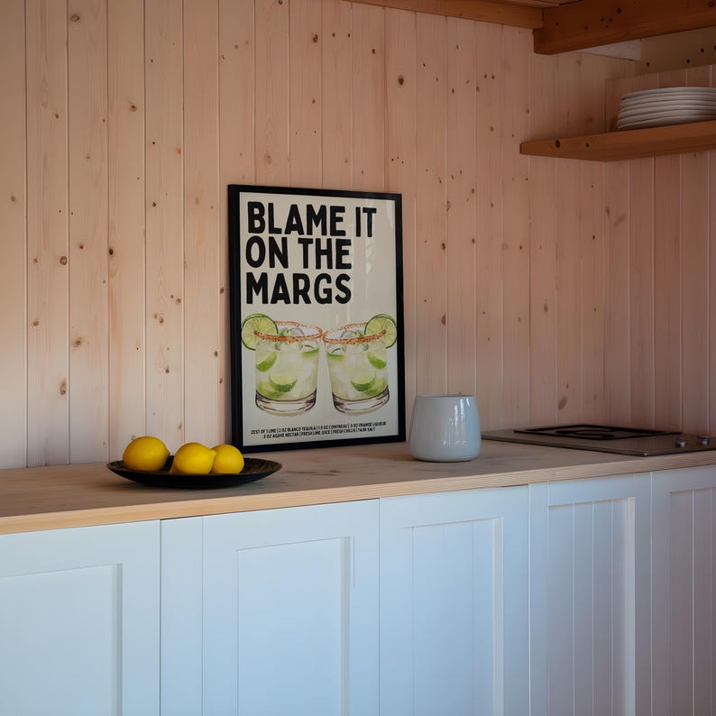 May include: A kitchen scene with a framed poster that reads "BLAME IT ON THE MARGS" above a countertop. The poster features two margarita glasses. A black plate holds lemons, and a white vase sits nearby.