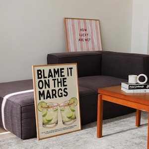 May include: Two framed art prints. One says "Blame it on the Margs" with margarita illustrations. The other says "How lucky are we?" with pink and white stripes. Both are in wooden frames.
