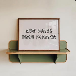 May include: A framed white print with the text "SAVE WATER DRINK MONSTER" in black, displayed on a green and wood-toned shelf. The shelf is mounted on a white wall, creating a minimalist aesthetic.