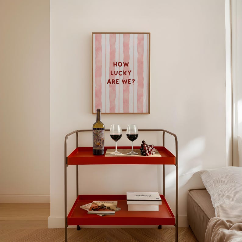 May include: A red metal bar cart with two shelves, a bottle of wine, two wine glasses, and a bowl of grapes. The cart is in front of a white wall with a framed print that says "How lucky are we?"