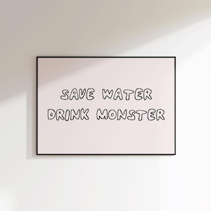 May include: A framed print with the text "SAVE WATER DRINK MONSTER" in black, handwritten-style lettering on a light pink background. The frame is black. The print is displayed on a white wall.