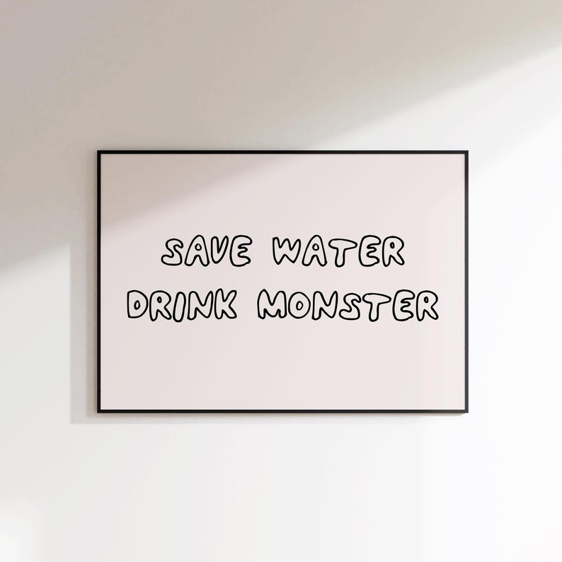 May include: A framed print with the text "SAVE WATER DRINK MONSTER" in black, handwritten-style lettering on a light pink background. The frame is black. The print is displayed on a white wall.