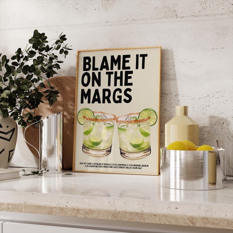 May include: A framed print with the text "BLAME IT ON THE MARGS" above an illustration of two margaritas. The print includes a recipe with measurements in ounces. The image is set on a kitchen counter with decorative items.