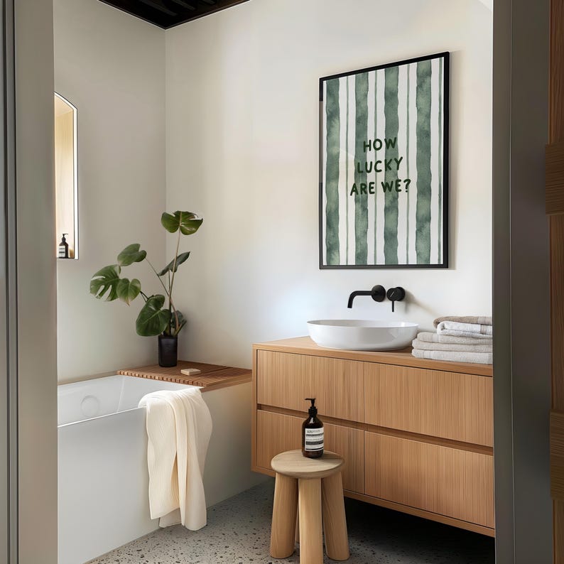 May include: A bathroom with a white bathtub, a wooden vanity with drawers, a white sink, and a framed print with green stripes and the text "How lucky are we?" hanging on the wall.