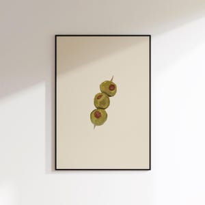 May include: A framed wall art print featuring a vertical illustration of three green olives with red pimento centres skewered on a thin wooden toothpick, centred against a solid light beige background within a thin black rectangular frame.