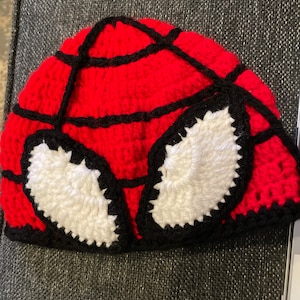 May include: Red and black crocheted hat with white crocheted eyes, resembling a superhero character.