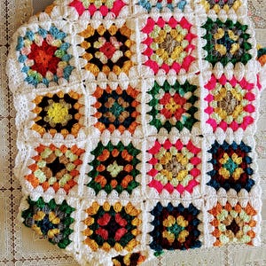May include: A colorful, handmade crochet blanket featuring a patchwork design. The blanket is composed of numerous square motifs in various colors, including red, yellow, green, and blue, all bordered by white yarn. The blanket is a cozy, decorative item.