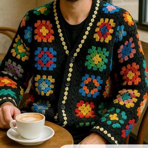 May include: Black crochet cardigan with colorful floral squares. The cardigan has a V-neck, button closure, and a cream-colored trim. A white coffee cup and saucer sit on a wooden table.