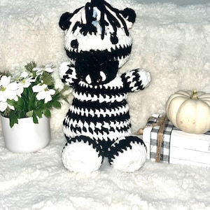 May include: A handmade, black and white crocheted zebra plush toy. The zebra has a striped body, a black mane, and white feet. It is sitting next to a white pumpkin, a small stack of books, and a vase of white flowers.