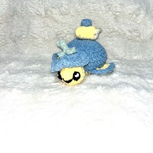 以下が含まれることがあります： 手作りの、漫画のキャラクターの形をしたかぎ針編みの玩具。おもちゃは主に青色で、黄色と薄緑色のアクセントがあります。笑顔の顔をしており、白いテクスチャード加工の背景に置かれています。
