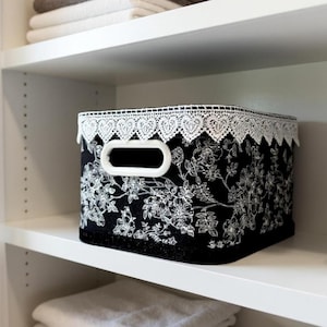 May include: Black storage bin with white floral pattern and a white lace trim. The bin has a white handle and is sitting on a white shelf. Several folded towels are stacked on the shelves.