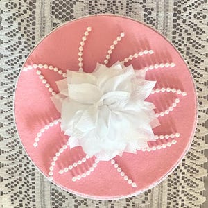 May include: A round, pink decorative box with a white, paper flower in the center. White pearl beads are arranged in a sunburst pattern around the flower. The box is on a white lace surface.
