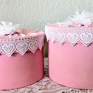 May include: Two pink round boxes with white lace trim featuring heart-shaped cutouts. Each box is topped with a white flower and faux pearls. The boxes are displayed on a white lace tablecloth.