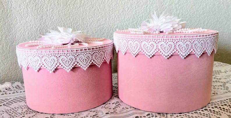 May include: Two pink round boxes with white lace trim featuring heart-shaped cutouts. Each box is topped with a white flower and faux pearls. The boxes are displayed on a white lace tablecloth.
