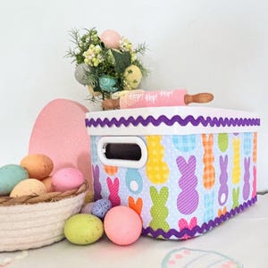 May include: A decorative Easter-themed storage box with a white handle and a colorful bunny pattern. The box is decorated with a purple scalloped trim. A basket of colorful eggs and a pink egg-shaped decoration are also present.