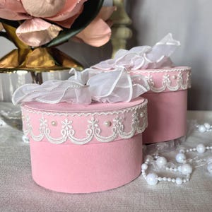 May include: Two pink, round boxes with white lace trim and pearl accents. Each box is topped with a white, ruffled fabric. The boxes are on a light-colored surface, with a string of pearls in the background.