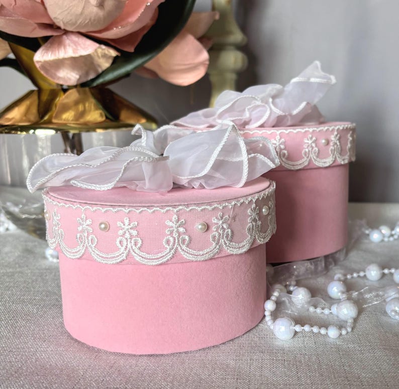 May include: Two pink, round boxes with white lace trim and pearl accents. Each box is topped with a white, ruffled fabric. The boxes are on a light-colored surface, with a string of pearls in the background.