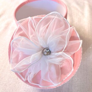 May include: A pink round gift box with a white and pink flower on top. The flower has a rhinestone center and white trim. The box is open, revealing a white interior.
