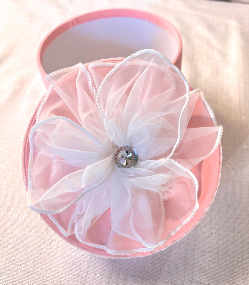May include: A pink round gift box with a white and pink flower on top. The flower has a rhinestone center and white trim. The box is open, revealing a white interior.