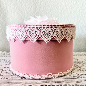May include: A round, pink box with white lace trim featuring heart-shaped cutouts. The box is topped with a white flower and sits on a white lace tablecloth. The box is a decorative item.