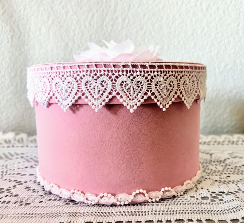 May include: A round, pink box with white lace trim featuring heart-shaped cutouts. The box is topped with a white flower and sits on a white lace tablecloth. The box is a decorative item.
