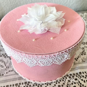 May include: A pink round box with a white lace trim and a white fabric flower on top. Small pearl beads are scattered on the lid. The box is likely for storage or decoration.