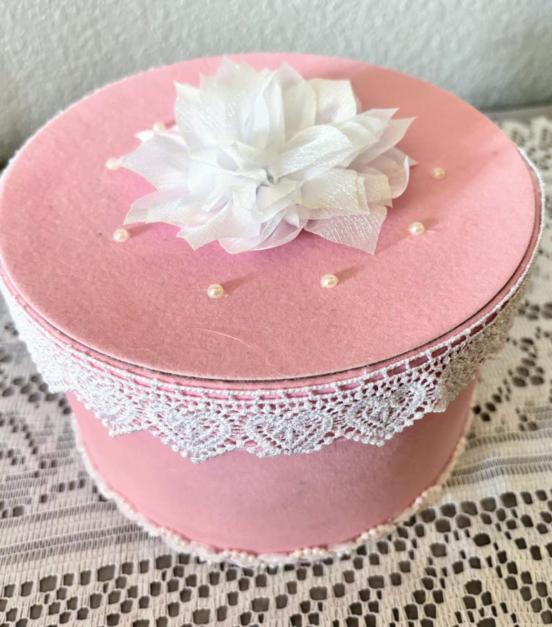 May include: A pink round box with a white lace trim and a white fabric flower on top. Small pearl beads are scattered on the lid. The box is likely for storage or decoration.