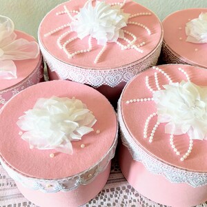May include: A collection of pink, round boxes with white lace trim and decorative white fabric flowers on top. Some boxes have pearl accents. The boxes are arranged on a white lace tablecloth.