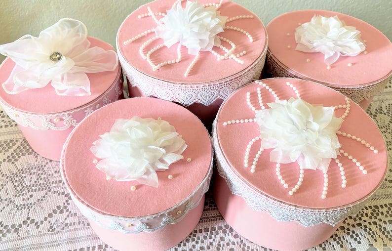 May include: A collection of pink, round boxes with white lace trim and decorative white fabric flowers on top. Some boxes have pearl accents. The boxes are arranged on a white lace tablecloth.