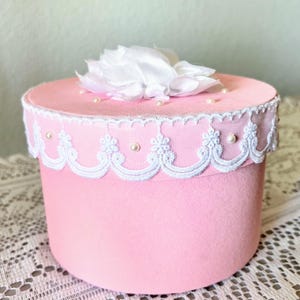May include: A pink, cylindrical box with a white lace trim and a white fabric flower on top. Small pearls are scattered on the lid. The box is likely for storage or decoration.