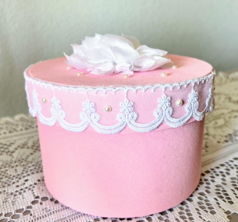 May include: A pink, cylindrical box with a white lace trim and a white fabric flower on top. Small pearls are scattered on the lid. The box is likely for storage or decoration.