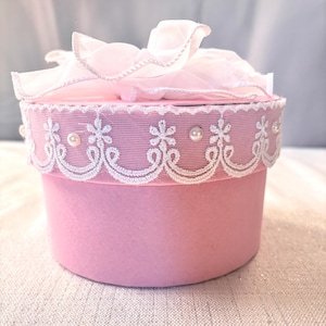 May include: A round, pink gift box with a white lace trim and pearl accents. The lid is topped with a delicate, ruffled pink bow. The box is a soft, pastel pink color, perfect for a special occasion.