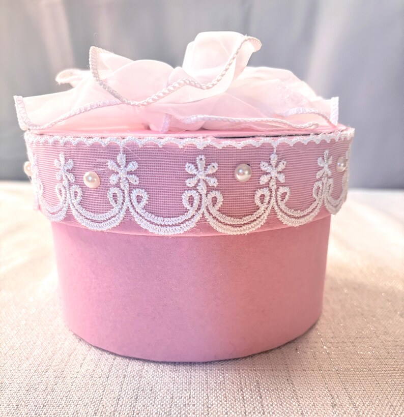 May include: A round, pink gift box with a white lace trim and pearl accents. The lid is topped with a delicate, ruffled pink bow. The box is a soft, pastel pink color, perfect for a special occasion.