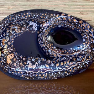 以下が含まれることがあります： 金と白の模様が施された黒い楕円形のサービングディッシュ。ディッシュの中央には、小さくて丸いディップボウルがあります。デザインには、スタイリッシュな葉、星、点々が描かれています。