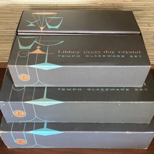 Può includere: Un set di quattro scatole contenenti bicchieri Libbey Tempo. Le scatole sono grigie con un design blu e oro. La scatola superiore riporta la scritta "Libbey every-day crystal TEMPO GLASSWARE SET".