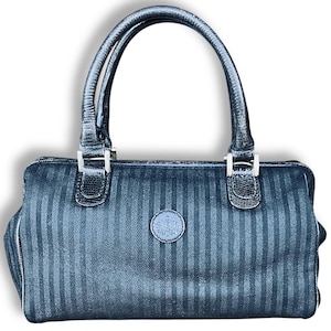 May include: A black handbag with vertical stripes and a rounded shape. The bag has two handles and silver-colored hardware. A small, circular emblem is centered on the front. The bag's design features a textured, patterned surface.