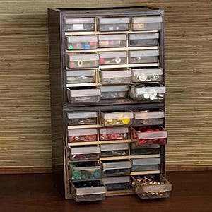 Może przedstawiać: Czarny plastikowy organizer szufladkowy z wieloma małymi szufladami wypełnionymi różnymi guzikami, koralikami i innymi małymi materiałami do rękodzieła.