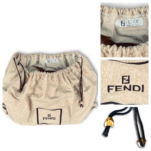May include: Beige Fendi dust bag with a drawstring closure. The bag features the Fendi logo in a black rectangle on the front. The drawstring is black with gold-colored metal tips. The bag is made of a soft, textured fabric.