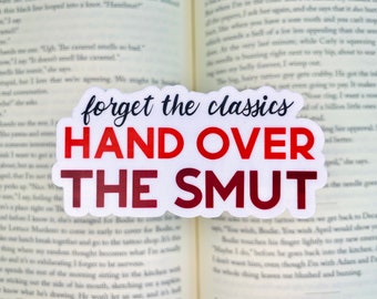 May include: A white sticker with the text "forget the classics HAND OVER THE SMUT" in a mix of red and black fonts. The sticker is placed on top of an open book.