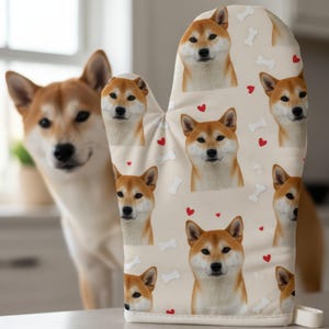 May include: A beige oven mitt featuring a repeating pattern of Shiba Inu dog faces, red hearts, and bone shapes. A real Shiba Inu dog is in the background. The oven mitt has a white loop for hanging.