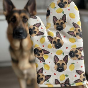 Op de afbeelding: Een witte ovenwant met een herhaald patroon van Duitse herdershondengezichten, gele citroenen en kleine rode hartjes. Op de achtergrond is een Duitse herdershond te zien.