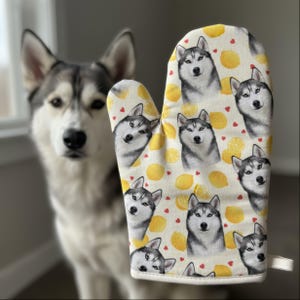 Puede incluir: Un guante de cocina blanco con un estampado de caras de Husky siberiano grises y blancos, limones amarillos y corazones rojos. El guante está hecho de tela suave y tiene un lazo para colgar. Un perro Husky está en el fondo.