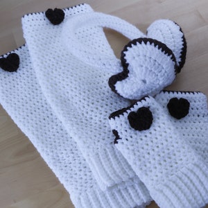 May include: A white crochet winter set with brown heart accents. The set includes a scarf, earmuffs, and fingerless gloves. The edges of the set are trimmed with brown yarn. The set is made of a chunky, textured yarn.