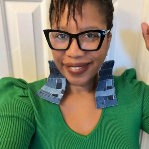 Könnte beinhalten: Eine Frau trägt einen grünen Pullover mit tiefem V-Ausschnitt und großen Ohrringen aus Jeans-Patchwork. Sie trägt eine schwarze Cat-Eye-Brille.