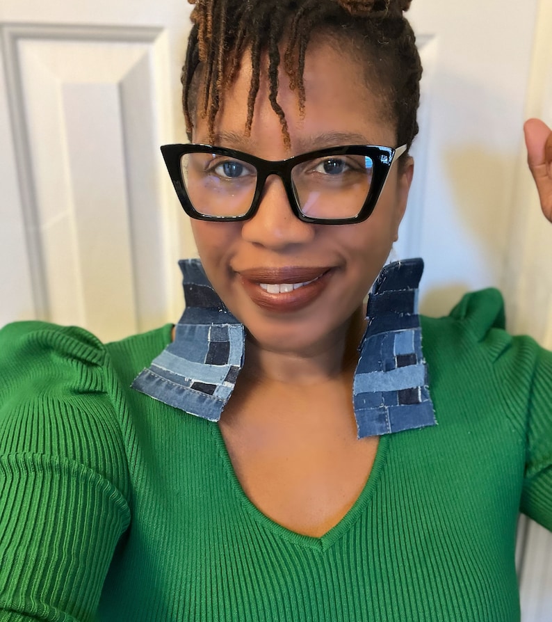 May include: A woman wearing a green sweater with a deep v-neck and large denim patchwork earrings. She is wearing black cat-eye glasses.