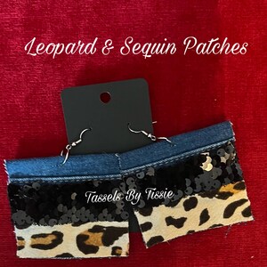 May include: A pair of denim earrings with black sequins and a leopard print fabric. The earrings are handmade and have a silver hook closure. The earrings are labeled "Tassels By Tisse".